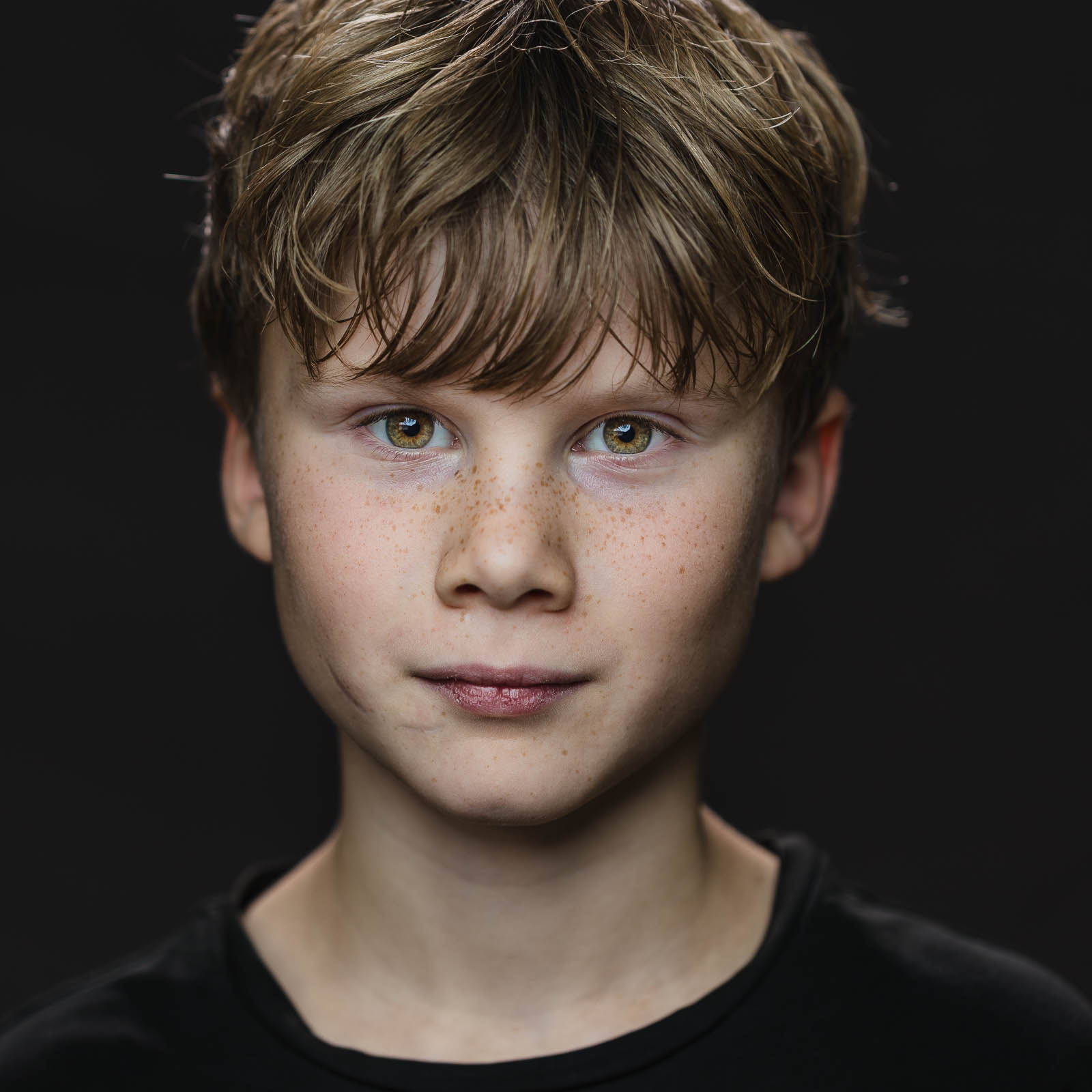 Fine art portret van een jonge jongen met lichte ogen, sproeten en nat haar, kijkend in de camera tegen een donkere achtergrond. Natuurlijke uitstraling en zachte belichting.